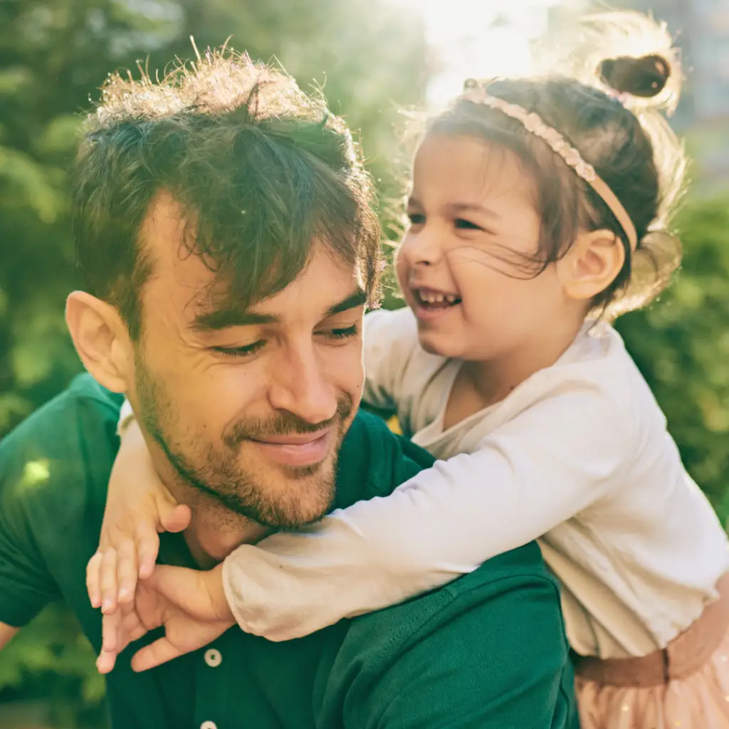 A little girl hugging her smiling father while sun shines between trees.