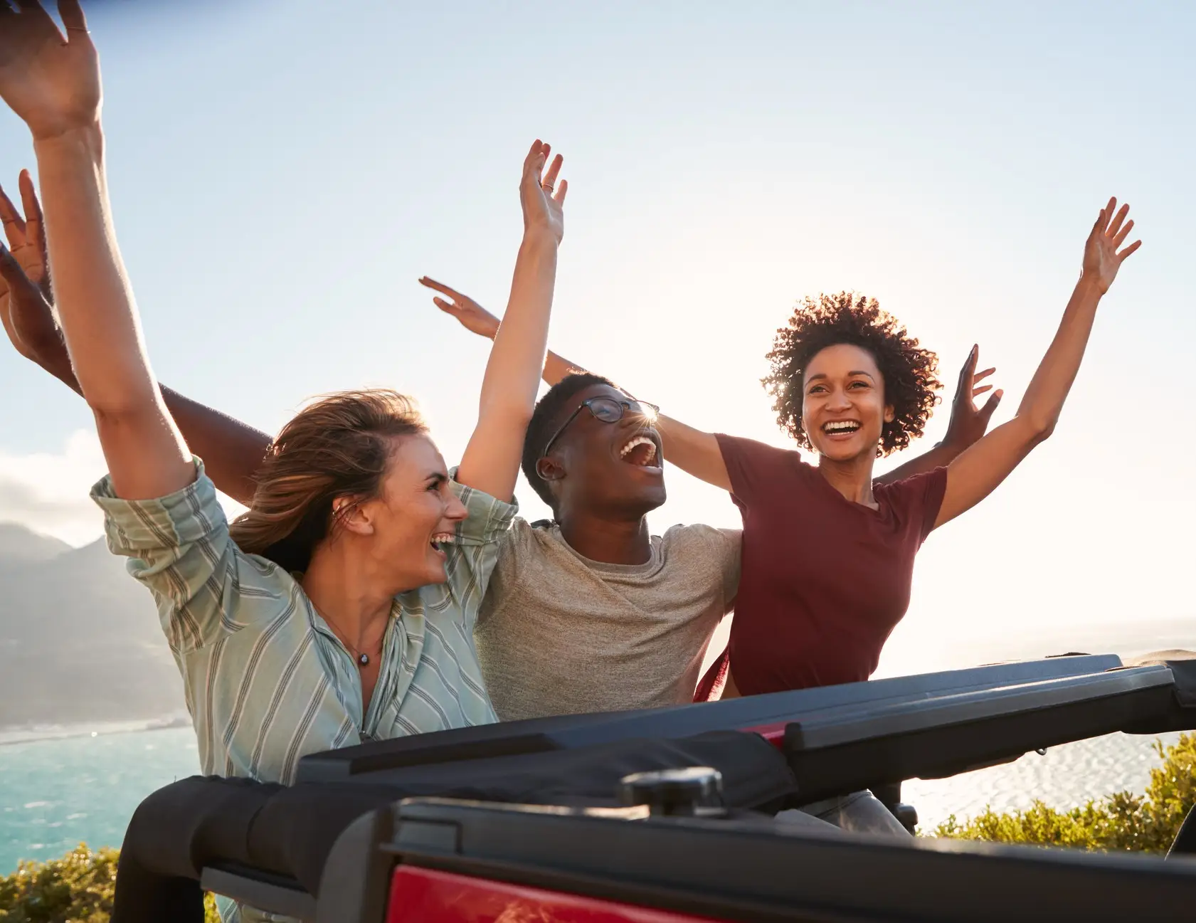 Excited millennial friends travelling in the back of an open car with their arms in the air