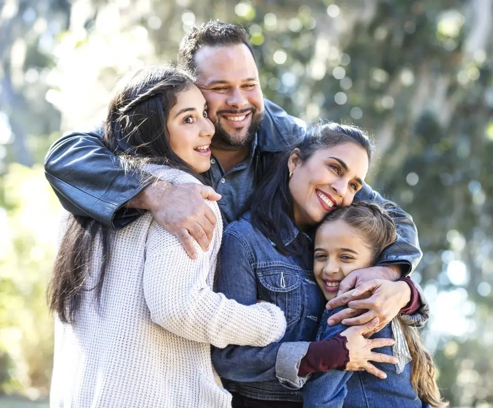 An Hispanic family of four, with two girls, standing outdoors at the park on a sunny autumn day, smiling, looking away from the camera and hugging one another. The younger daughter, 9 years old, is standing next to the mother. Her sister, a 13 year old teenager, is standing next to father.