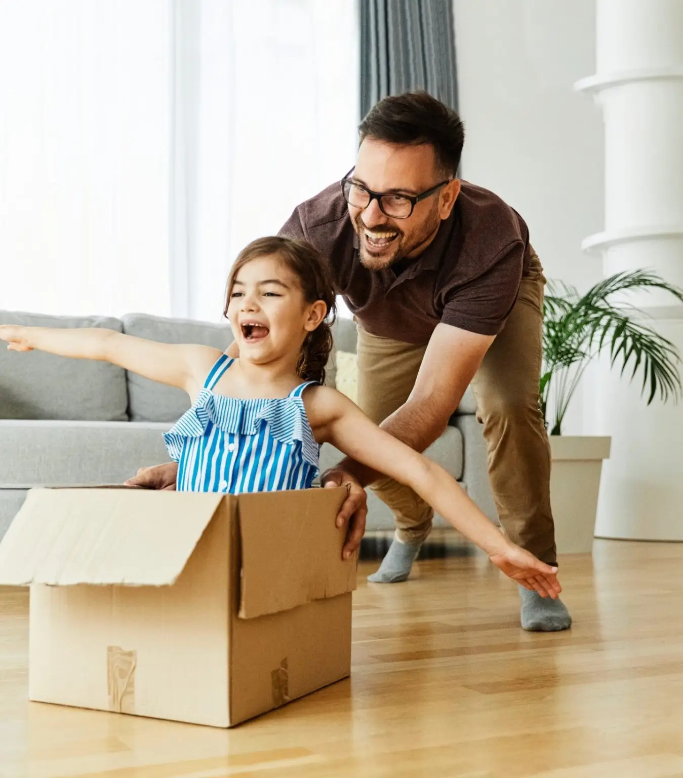 Father pushing his daughter sitting in a cardboard box, having fun at home