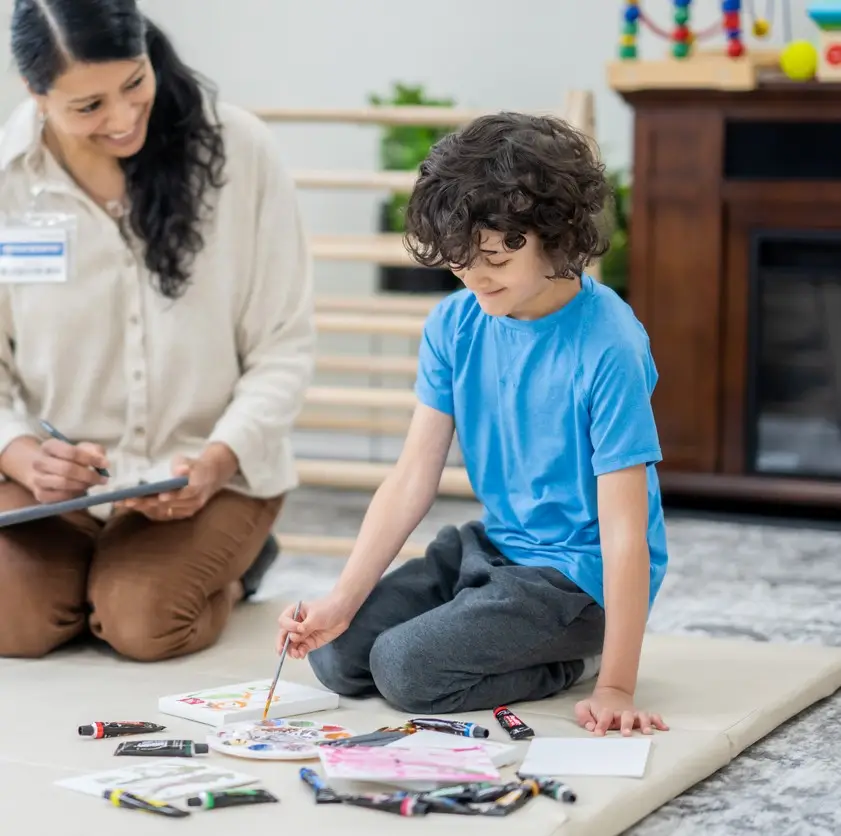 Child & Teen Therapy. A young boy lays out on the floor with various art supplies in front of him as he works with an Occupational Therapist. The Therapist is seated beside him and is holding a tablet as she takes notes during the appointment.