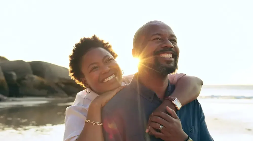 Couple, hug and happy on beach with love, bonding and sunset for holiday, vacation and relationship. Black people, man or woman with embrace, smile and romance by ocean or sea for travel or adventure