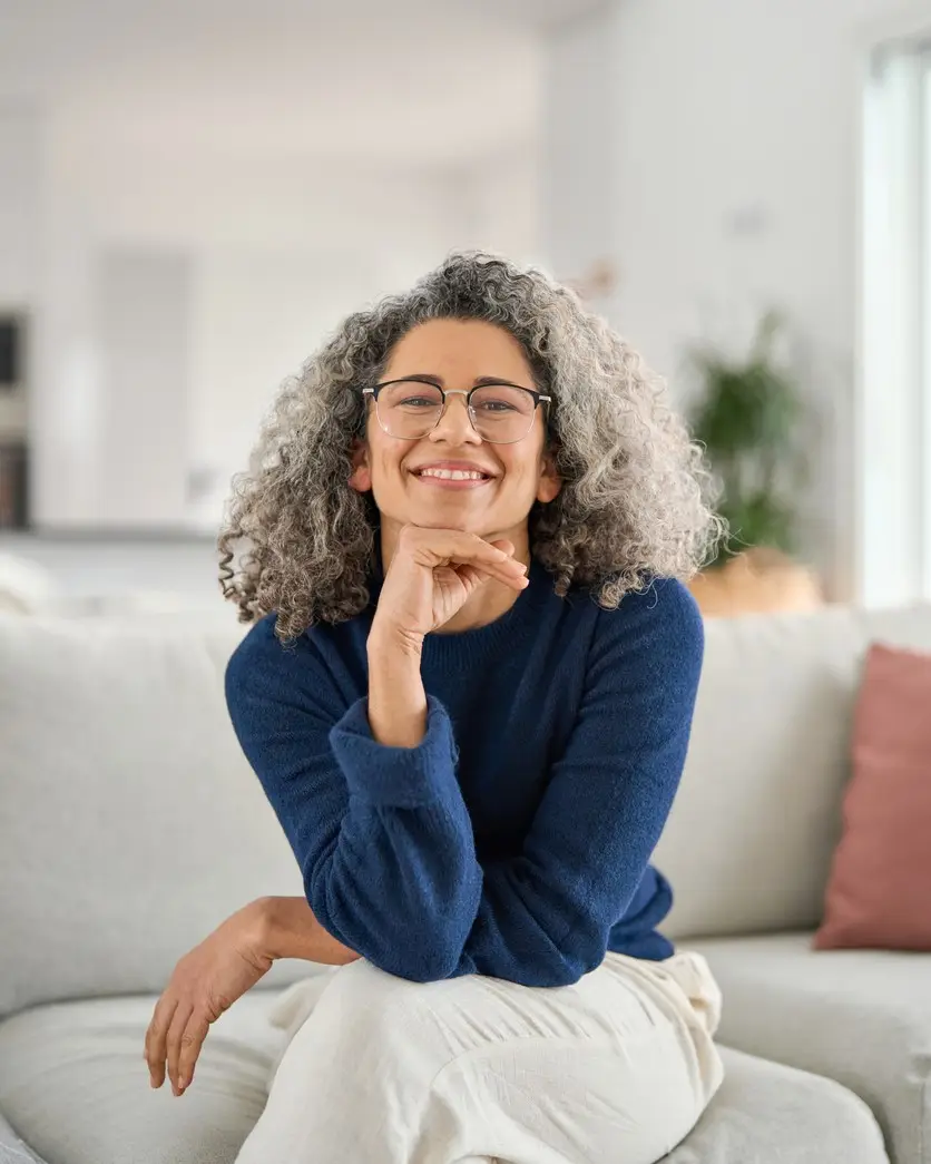 Vertical portrait of happy 50 years old woman sitting on sofa at home. Smiling middle aged lady looking at camera in living room at home. Mature older female with curly gray hair relaxing on couch.