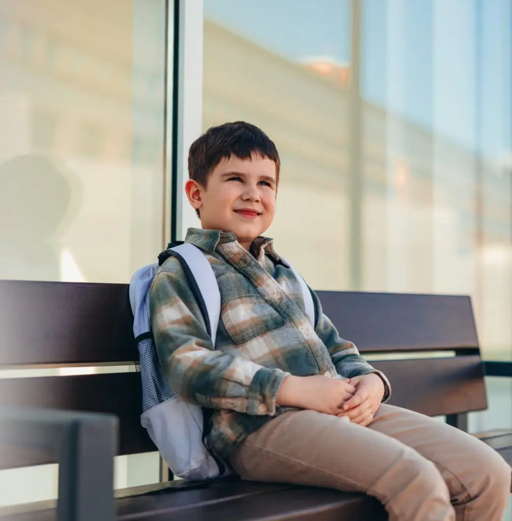 CBT Therapy. Elementary age boy sitting on a bench outdoors, waiting for school to start, looking away with a smile, embodying the joy and anticipation of childhood in a vibrant city setting