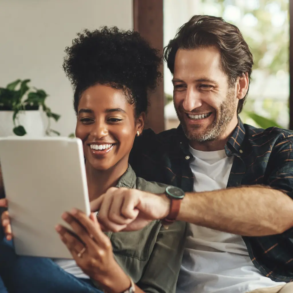 A couple sitting on a couch and looking at a tablet together while laughing