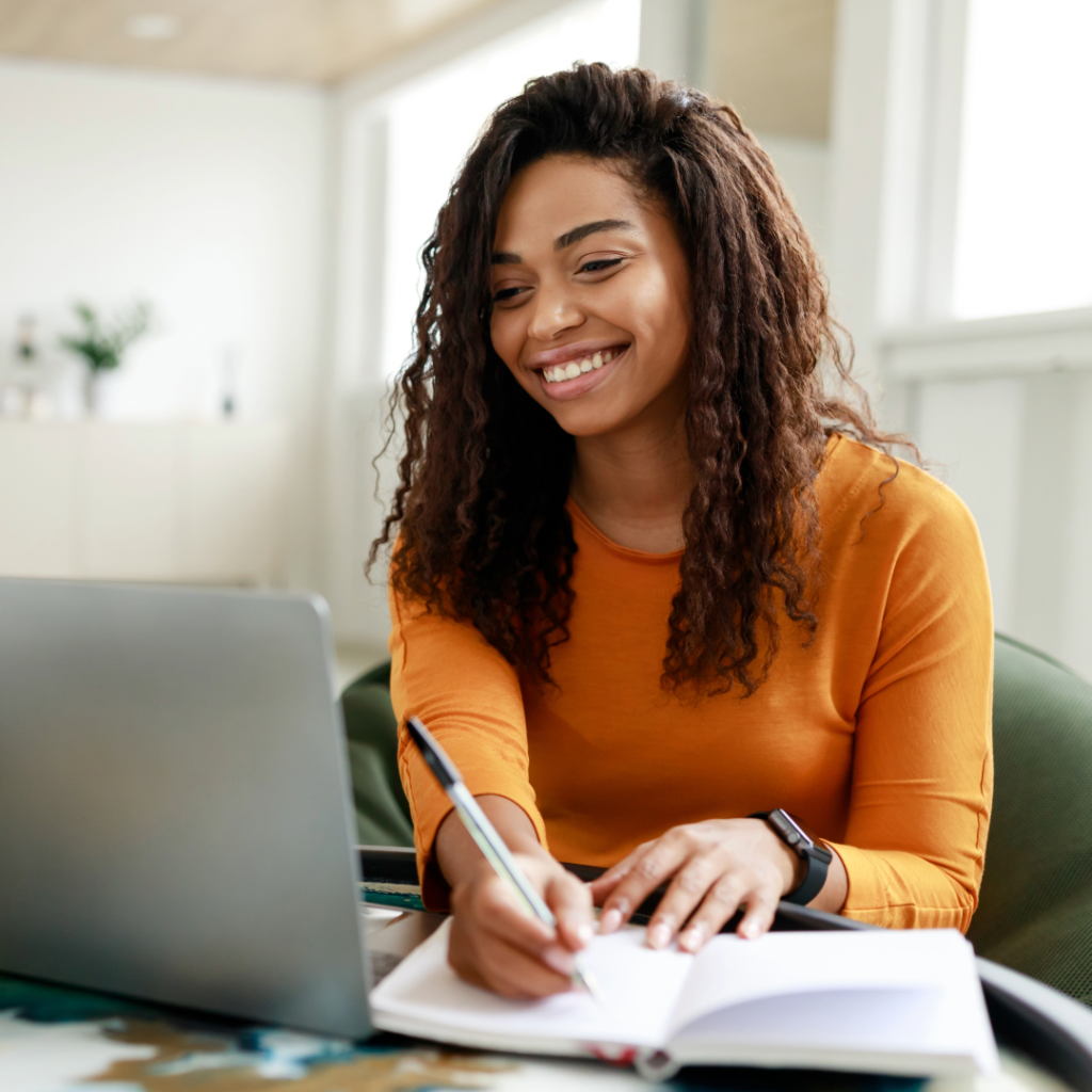DBT Therapy. Woman sitting at a kitchen table writing in notebook and looking at a laptop